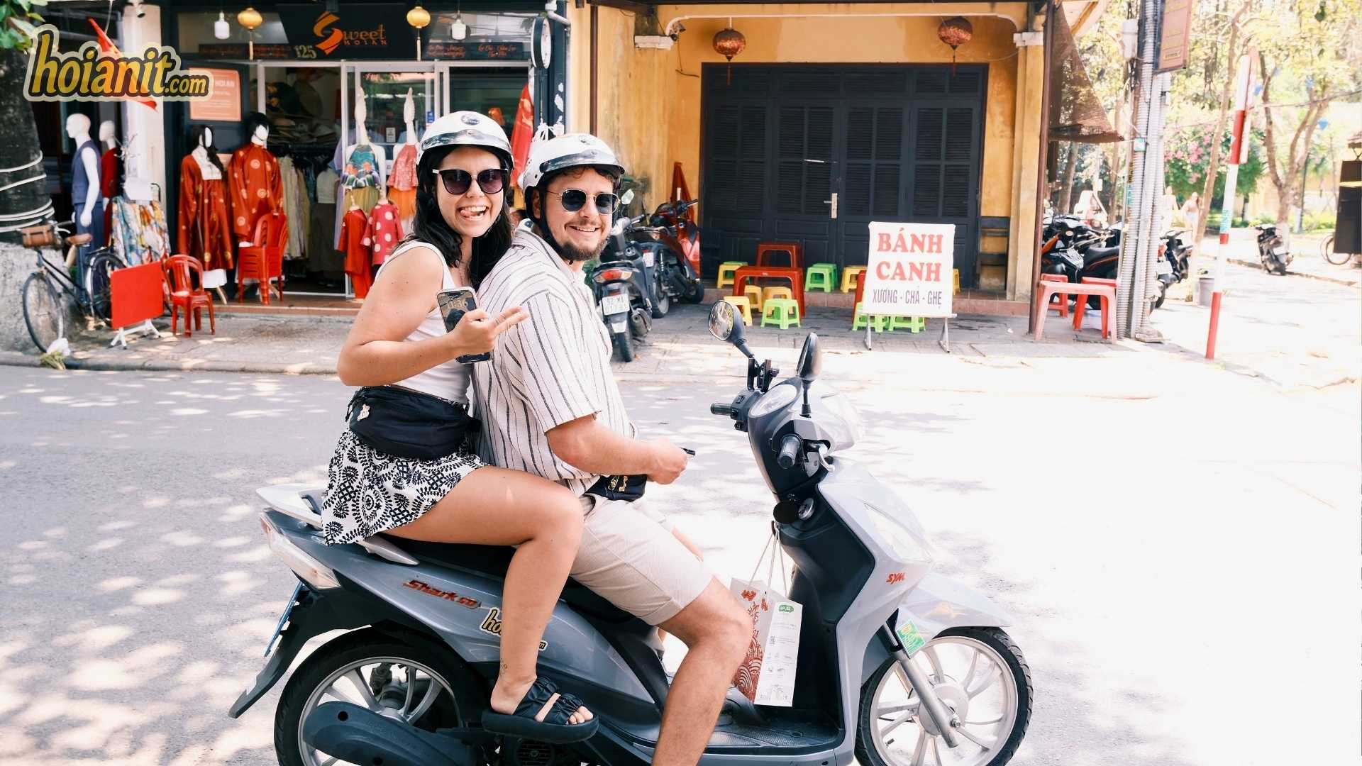Couple riding two-up on a small 50cc scooter in Hoi An, both wearing helmets