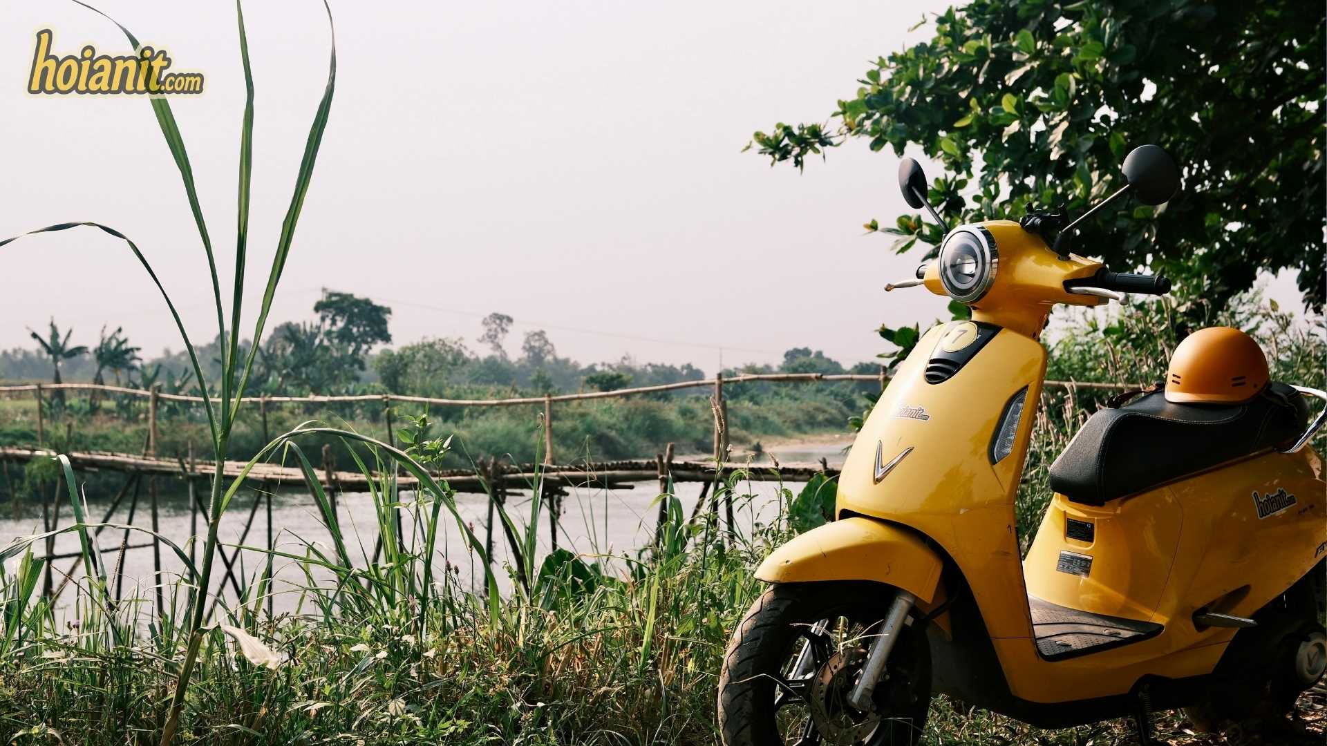 Yellow scooter parked by the river with the Cam Dong bamboo bridge in the distance