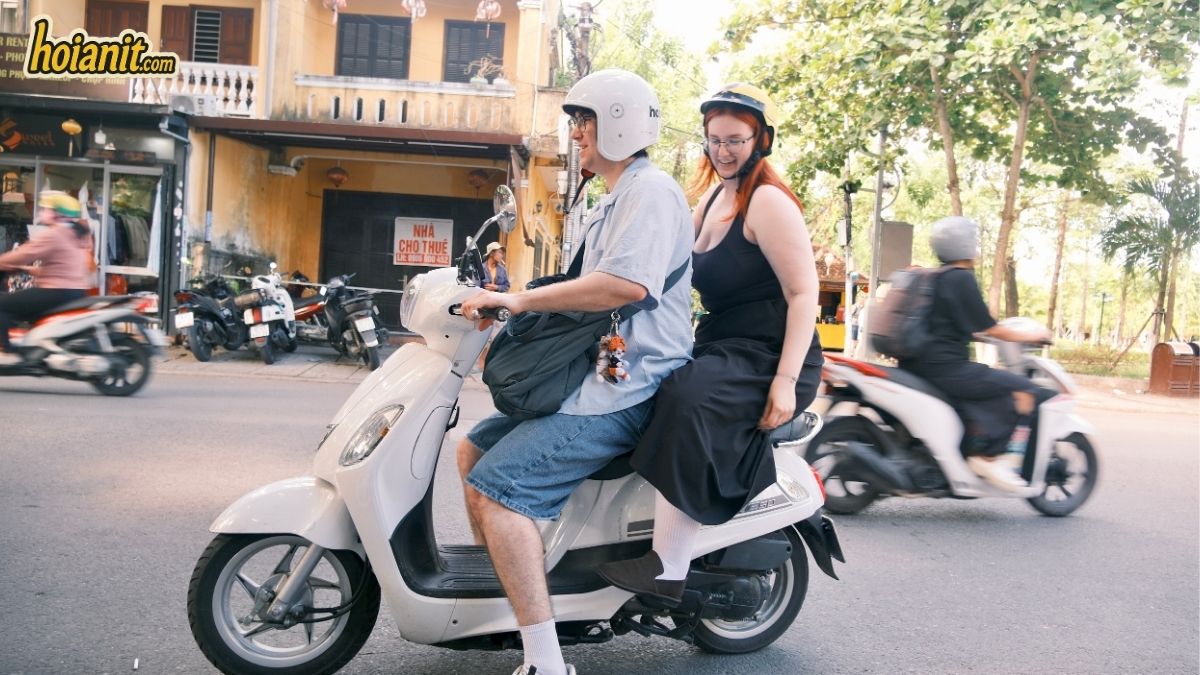 Pareja de turistas con cascos recorriendo el casco antiguo de Hoi An en scooter