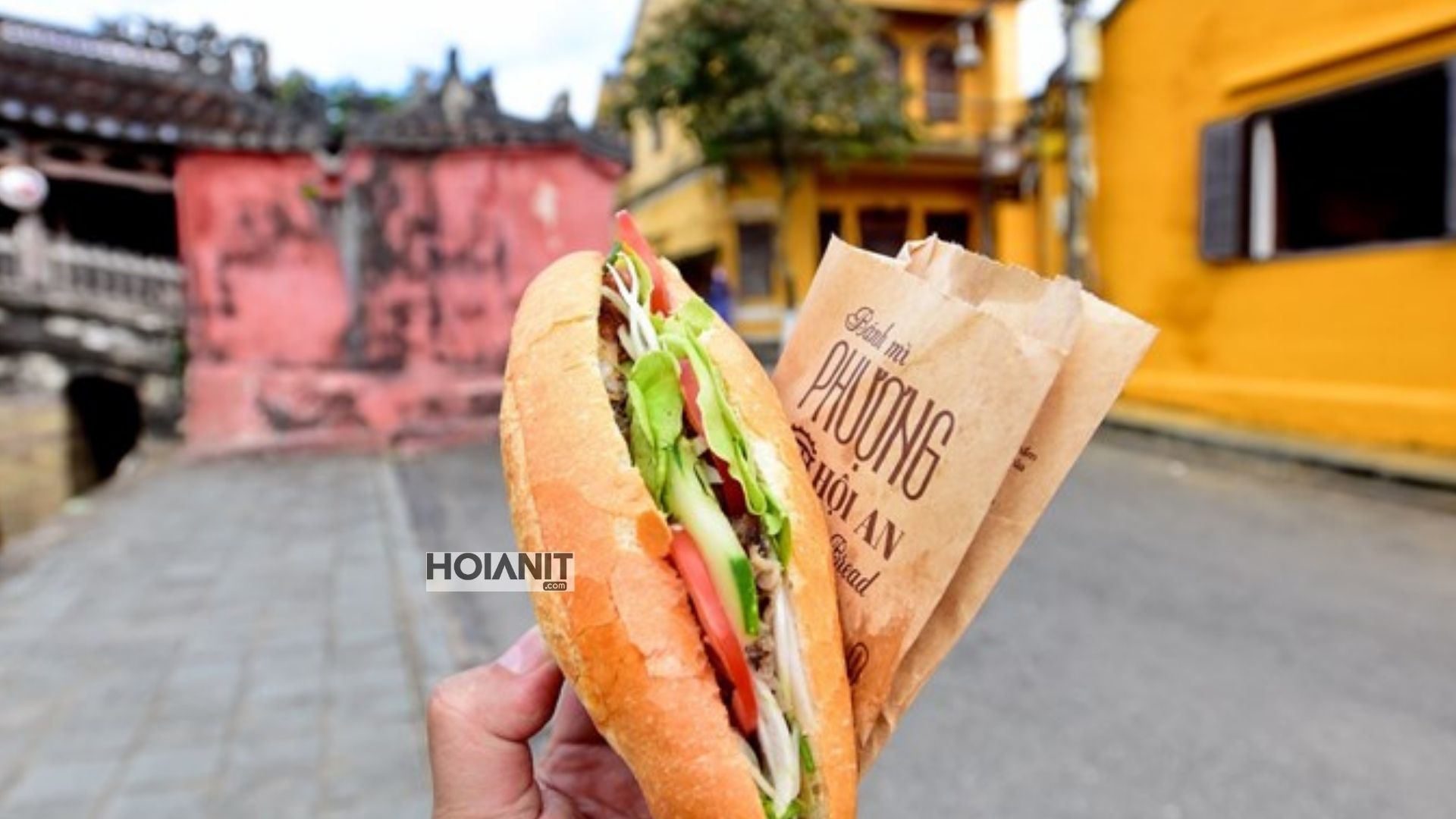 Ổ bánh mì Hội An gói giấy cầm trên tay trước bức tường vàng phố cổ