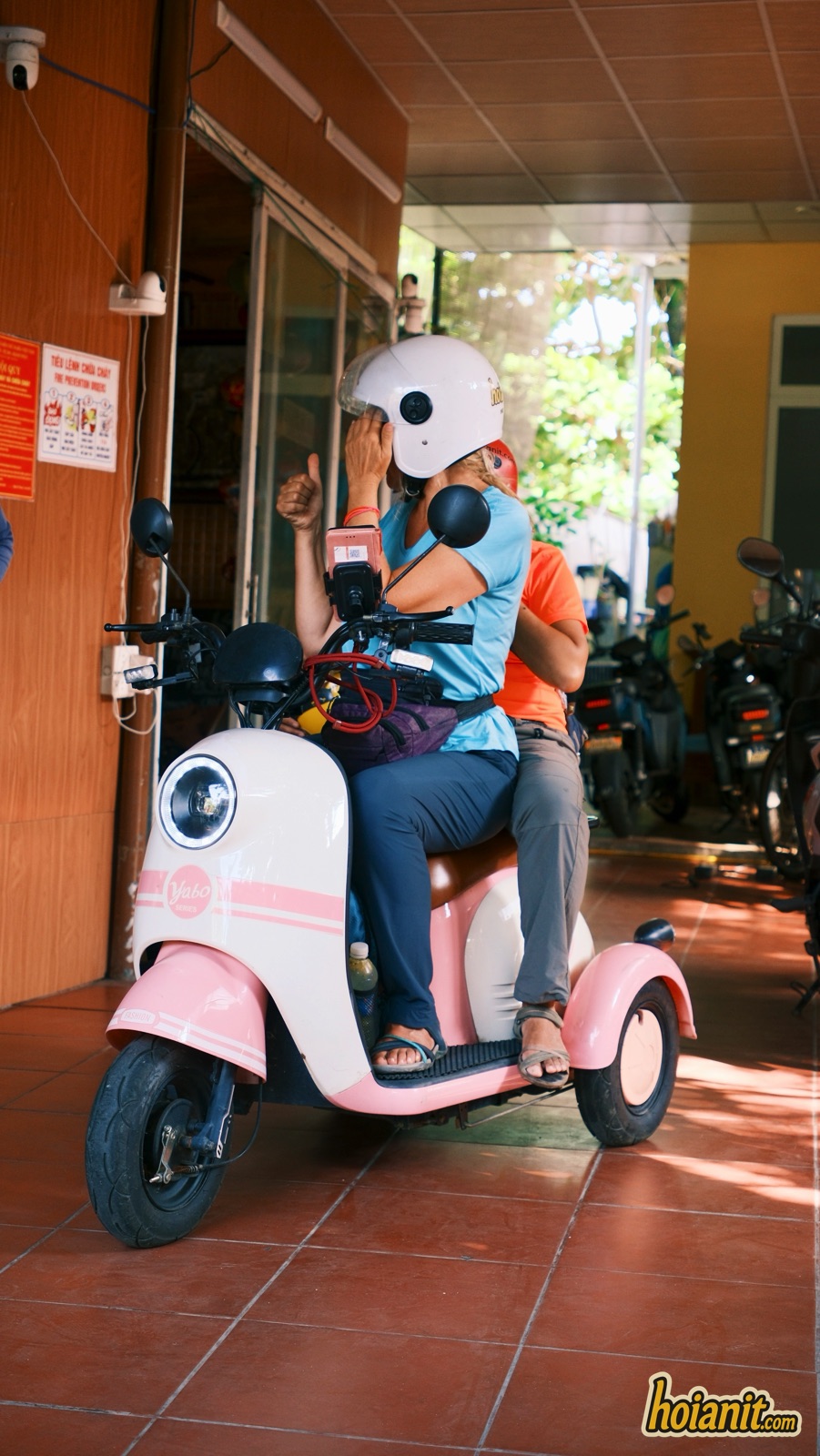Two riders on a small white and pink electric scooter at a Hoi An rental shop