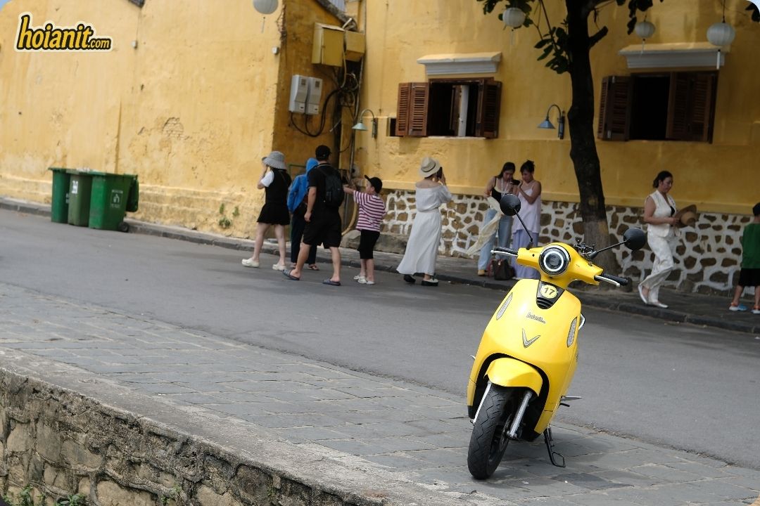 Yellow VinFast electric scooter parked by an ochre wall in Hoi An Old Town with tourists walking past