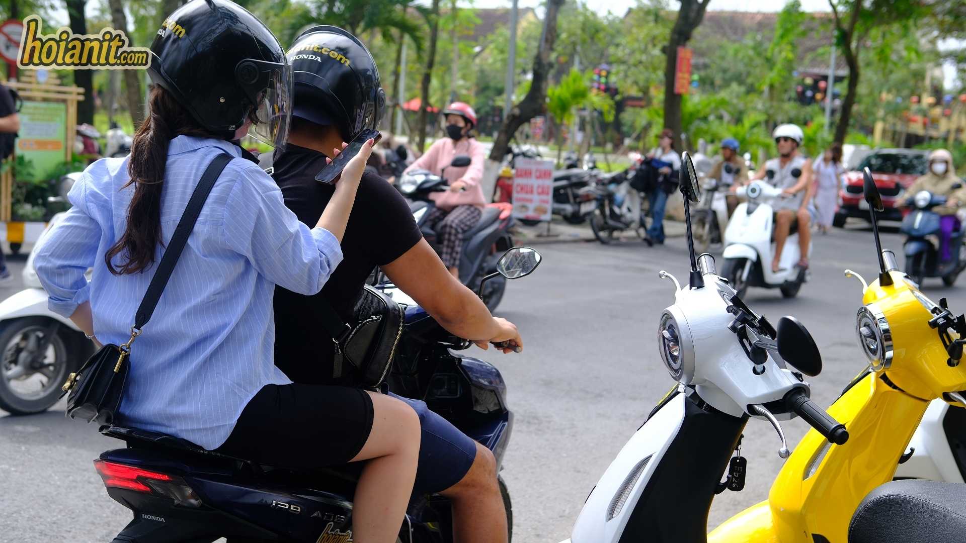 Two riders in helmets on a scooter on a park road in Hoi An