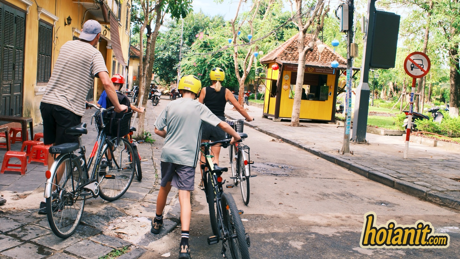 Family walking bicycles past a no-motorbike sign at a Hoi An Old Town entrance, where electric motorbikes are not allowed