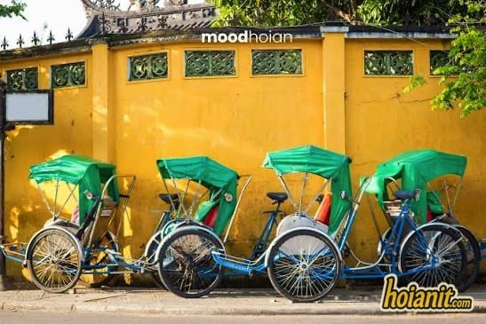 Bốn chiếc xích lô mui xanh đậu xếp hàng trước bức tường vàng đặc trưng Hội An